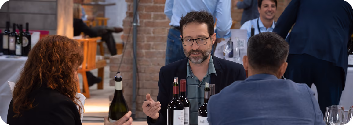 Three people seated at a table in a restaurant or wine bar, engaged in conversation, with bottles of red wine and wine glasses on the table.