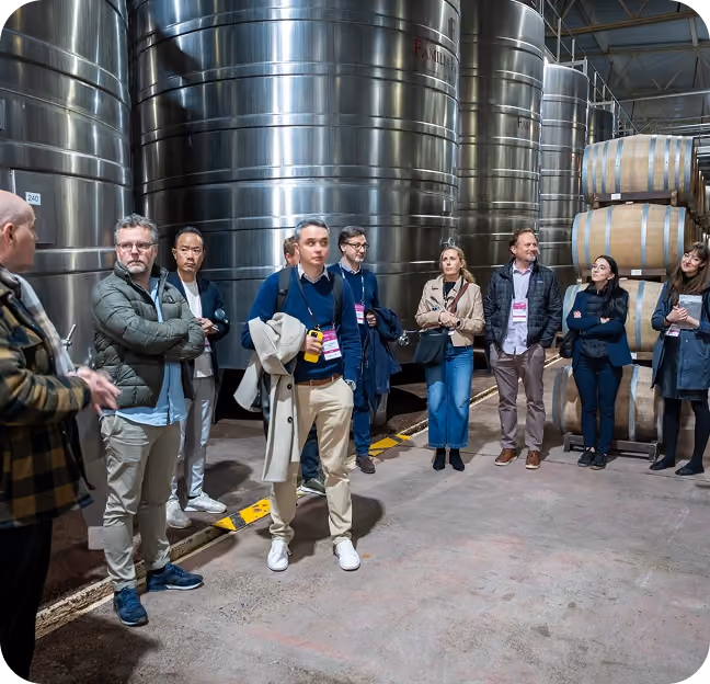 A group of people standing inside a large industrial facility with big metallic tanks and wooden barrels, listening to a man speaking on the left.