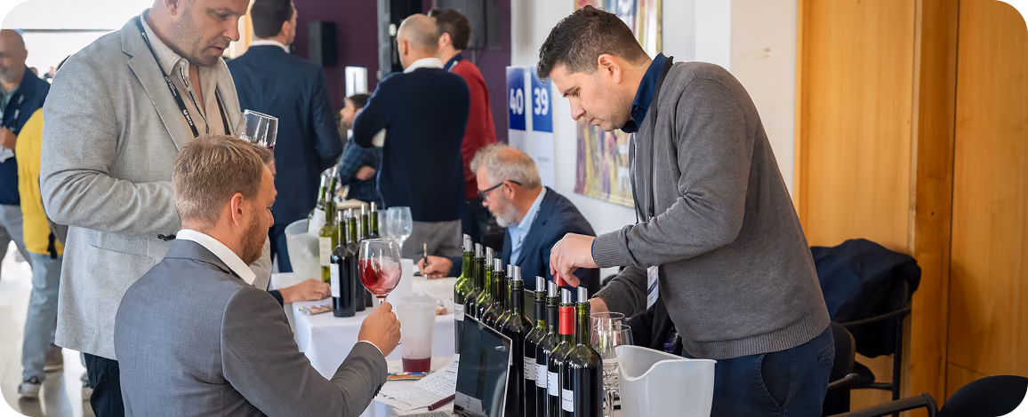 Two men at a wine tasting event; one pours wine from a bottle on a table lined with multiple wine bottles while the other holds a glass of red wine, with other attendees and tables visible in the background.