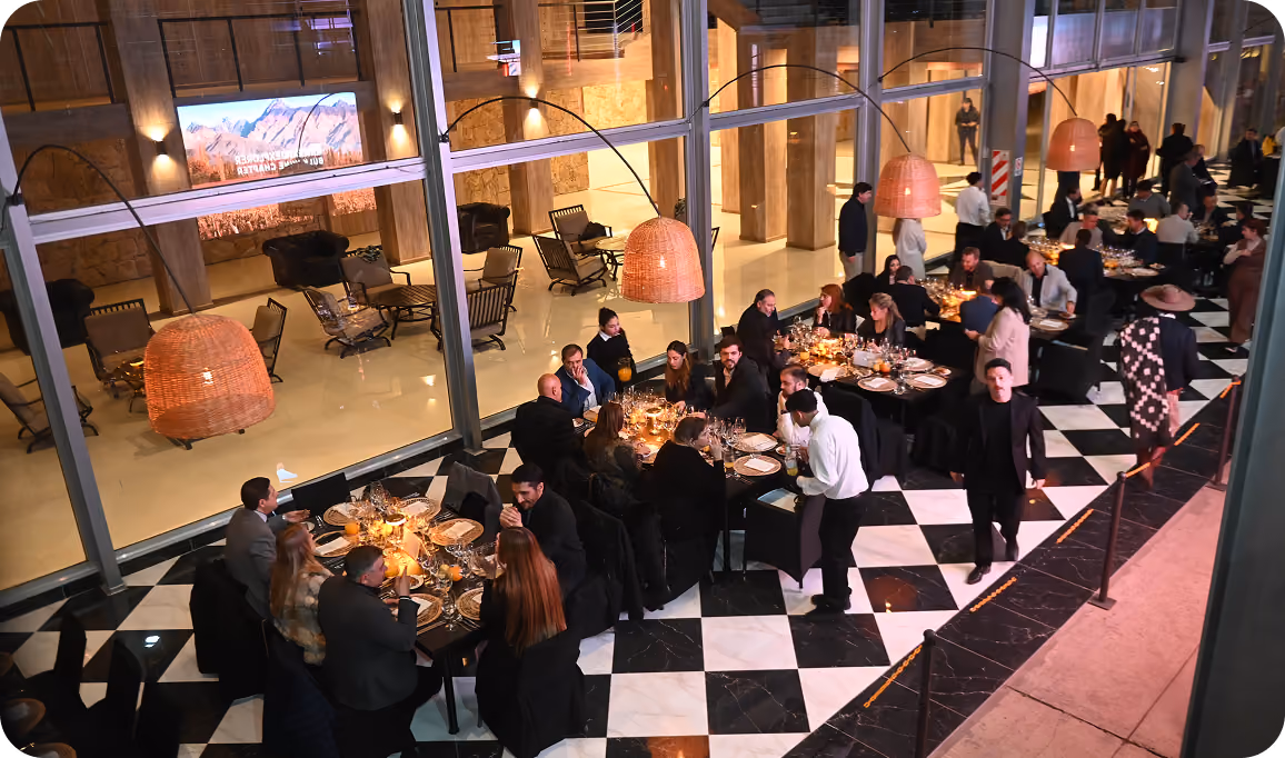 Evening indoor dining scene with multiple tables where people are seated, eating, and socializing under large hanging wicker lamps; the floor has a black and white checkered pattern and large glass windows separate the dining area from another seating space.