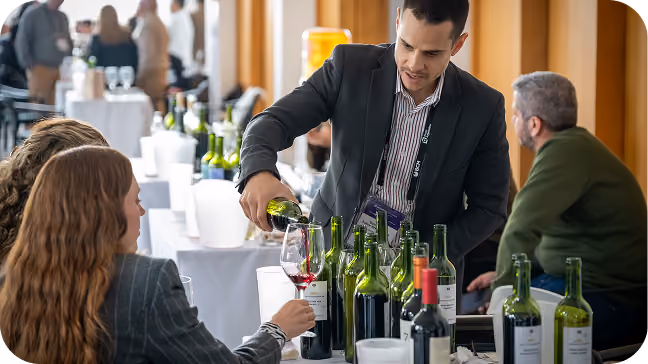 Man in a dark suit pouring red wine into a glass held by a seated woman, with several wine bottles on the table and people chatting in the background.