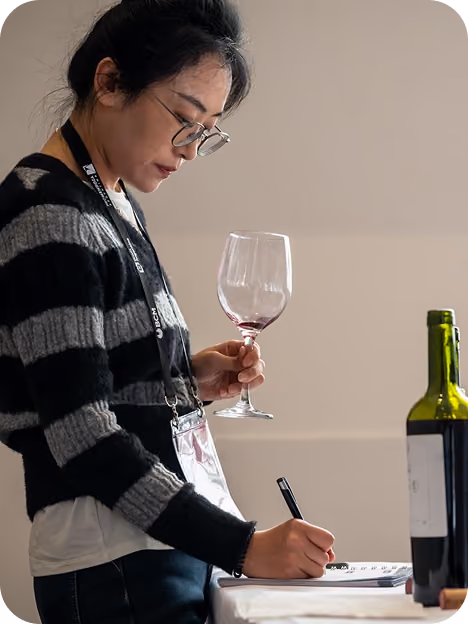Woman with glasses wearing a black and gray striped sweater holds a wine glass with red wine and writes notes on a paper at a table with an open wine bottle.