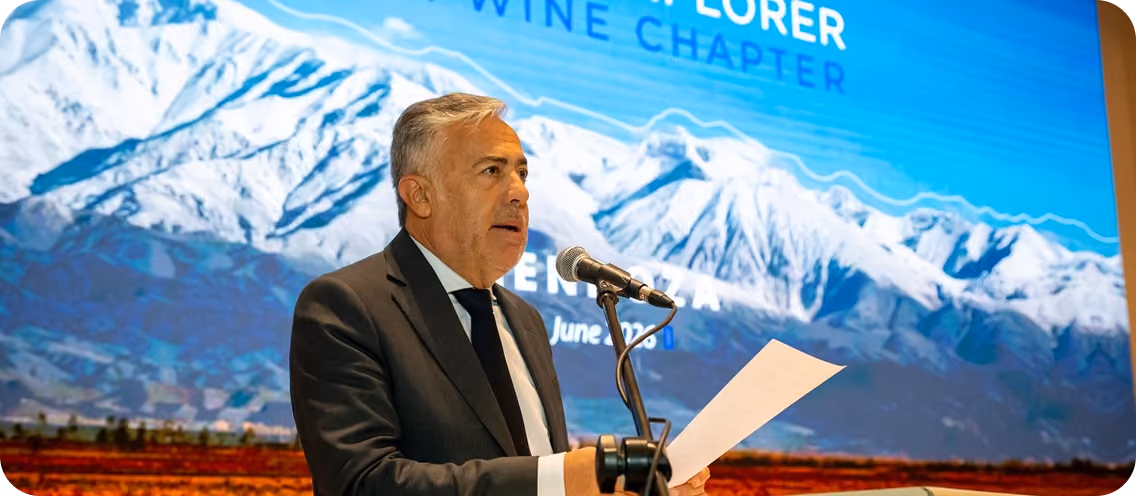 Man in a suit speaking into a microphone and holding a paper, standing in front of a large screen showing snowy mountains and text about a wine chapter event.