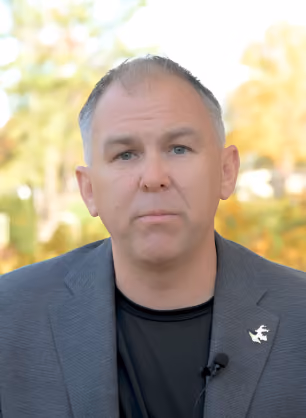 Man with short gray hair wearing a gray blazer over a black shirt, speaking in front of a blurred outdoor background with greenery and autumn colors.