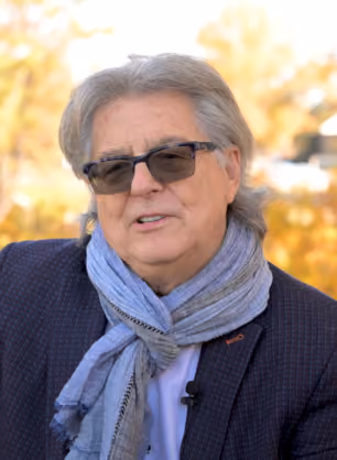 Older man with gray hair wearing dark sunglasses, a dark jacket, and a light blue scarf, with a blurred autumn background.