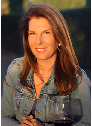 Woman with long brown hair wearing a denim jacket, smiling and holding a glass of red wine outdoors in natural light.