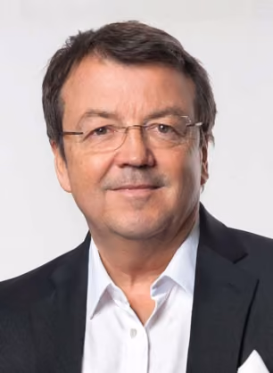 Middle-aged man with short dark hair and glasses, wearing a white shirt and dark blazer, looking directly at the camera with a slight smile on a plain light background.