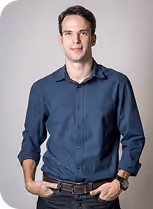 Portrait of a man with dark hair wearing a dark blue button-up shirt and brown belt, standing with hands in pockets against a plain light gray background.