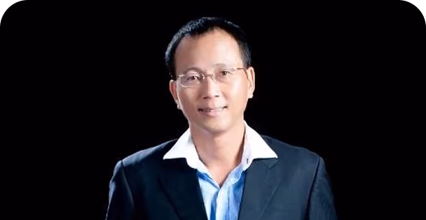 Man with short black hair and glasses wearing a dark suit jacket over a light blue shirt with a white collar, smiling against a black background.