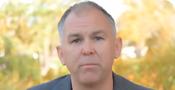 Middle-aged man with short gray hair and light complexion wearing a dark jacket, looking directly at the camera with a neutral expression, outdoors with blurred green and yellow foliage in the background.