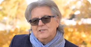 Older man with gray hair wearing dark sunglasses, a blue scarf, and a dark jacket, outdoors with a blurred background of autumn trees.