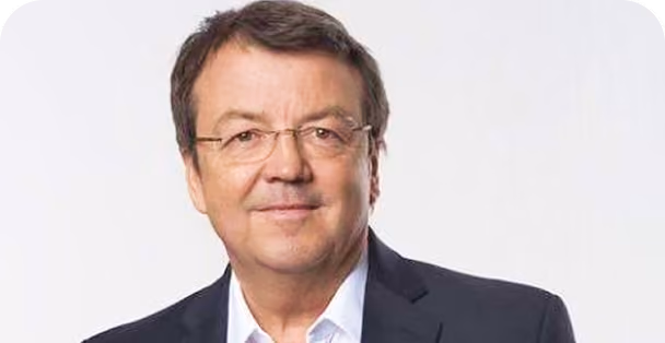 Portrait of a middle-aged man with short brown hair and glasses wearing a dark blazer and white shirt against a plain light background.
