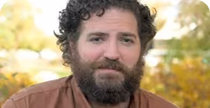 Man with curly dark hair, a full beard, and brown eyes wearing a brown shirt, outdoors with blurred greenery and sunlight in the background.