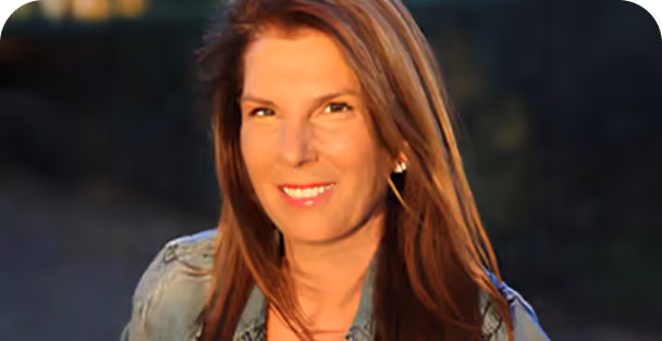 Woman with long brown hair smiling outdoors in soft natural light, wearing a denim jacket and pearl earrings.