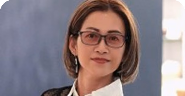 Portrait of a woman with short brown hair wearing glasses and a white collared shirt, standing against a blurred blue background.