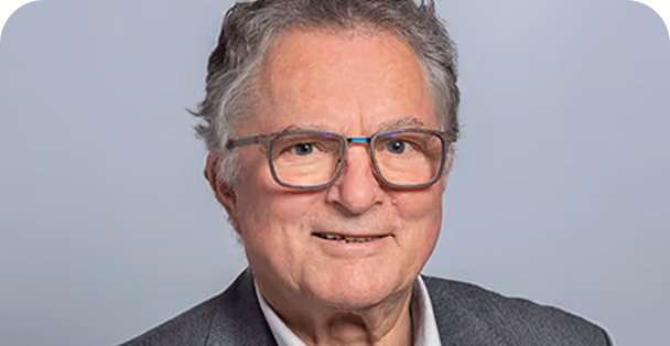 Portrait of an older man with grey hair and glasses wearing a dark blazer and light shirt, smiling against a plain light grey background.
