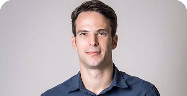 Portrait of a man with short dark hair wearing a dark blue button-up shirt, smiling slightly against a light gray background.