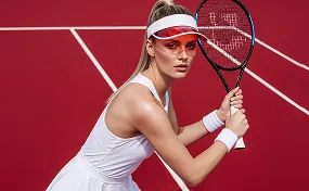 Female tennis player in white outfit and visor poised to hit a shot on a red tennis court with white lines.