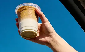 Hand holding up a clear plastic cup of iced layered coffee drink against a bright blue sky.