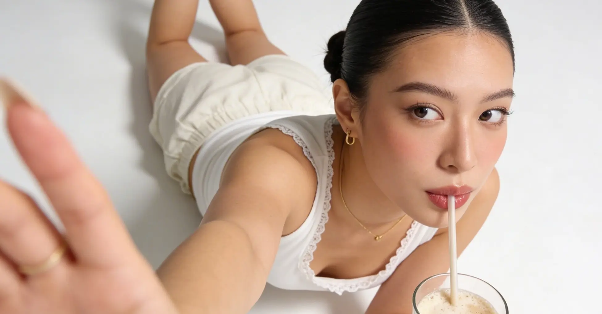 Young woman with dark hair tied back, wearing a white tank top and light shorts, lying on her stomach and drinking a frothy beverage through a straw while reaching forward.