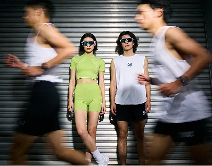 Two women standing against a metallic shutter background wearing sporty outfits and sunglasses, while two men in white tank tops and black shorts run past in a blur.