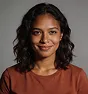 Portrait of a woman with medium-dark skin, curly hair, wearing a brown top, smiling gently against a dark background.