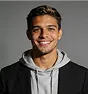 Smiling young man with short hair wearing a black blazer over a gray hoodie against a plain gray background.