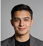Portrait of a young man with short dark hair wearing a dark gray blazer over a black shirt, smiling slightly against a plain light gray background.