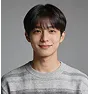 Portrait of a young man with short dark hair and a slight smile, wearing a gray striped sweater against a dark gray background.