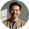 Smiling man with light brown skin and short curly black hair wearing a beige shirt against a light gray background.