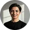 Smiling person with short dark hair wearing a black top, standing against a white wall with sunlight casting shadows.