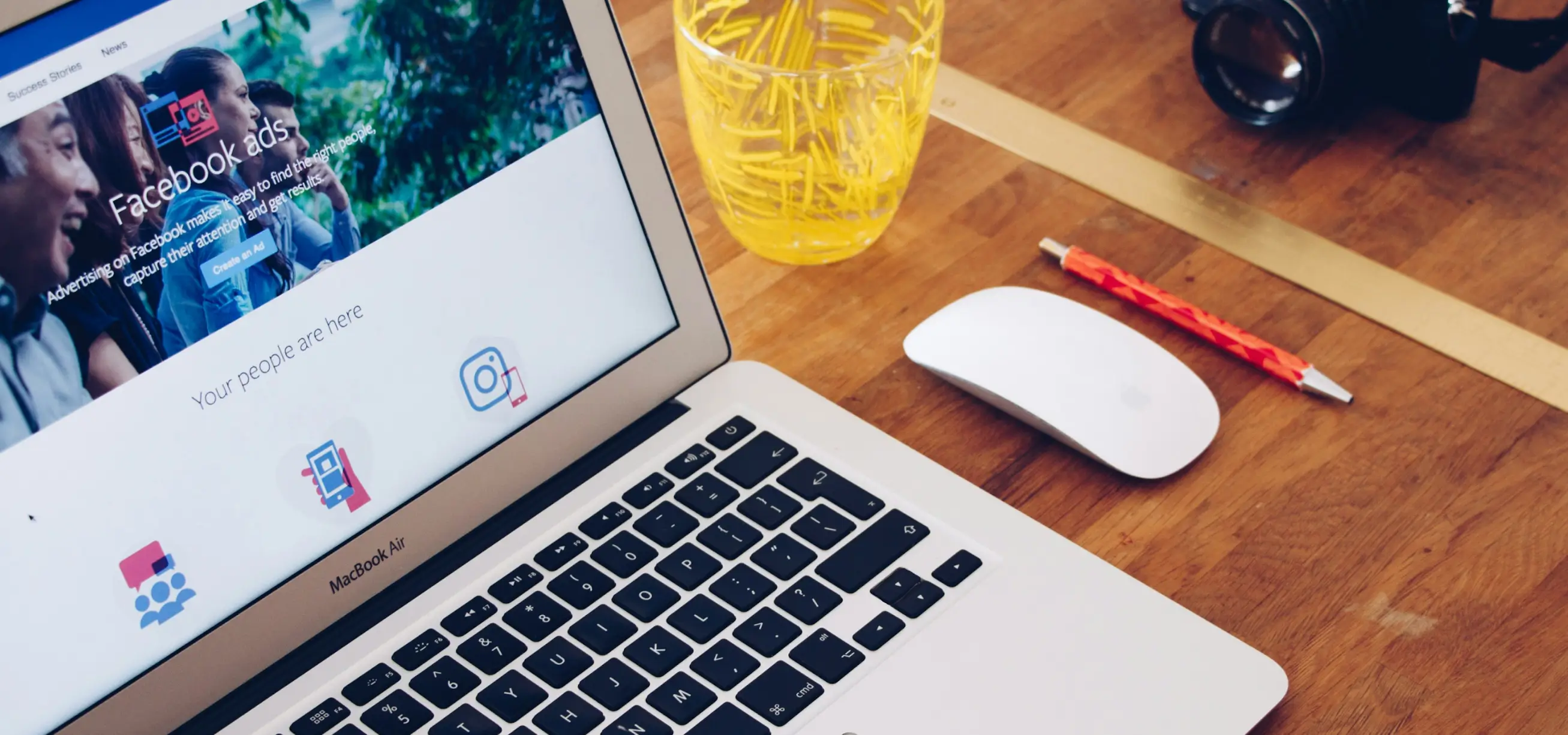 MacBook on a wooden desk showing the Facebook Ads homepage, next to a mouse and pens