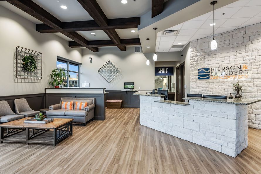 Modern lobby with a white stone reception desk, wooden ceiling beams, grey sofa, chairs, and wood-patterned flooring.