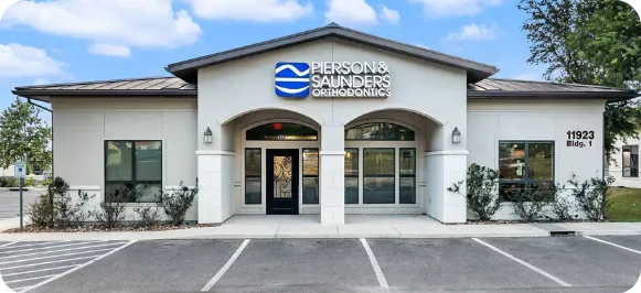 Single-story office building with Pierson & Saunders Orthodontics sign above the entrance, flanked by two large windows and surrounded by small shrubs, with an empty parking area in front.