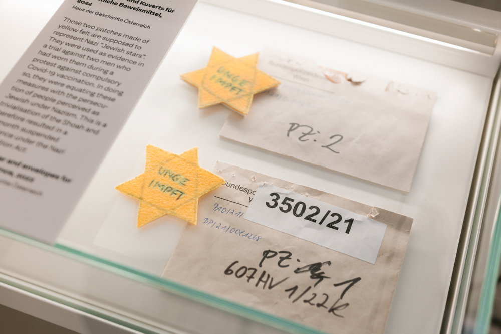 Zwei gelbe Filzsterne mit der Aufschrift „UNGEIMPFT“ neben beschrifteten Papierumschlägen in einer Vitrine des Hauses der Geschichte Österreich.