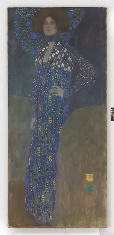 Gustav Klimt, Bildnis Emilie Flöge, 1902: Emilie Flöge in einem langen, blau gemusterten Kleid vor ornamentalem Hintergrund.