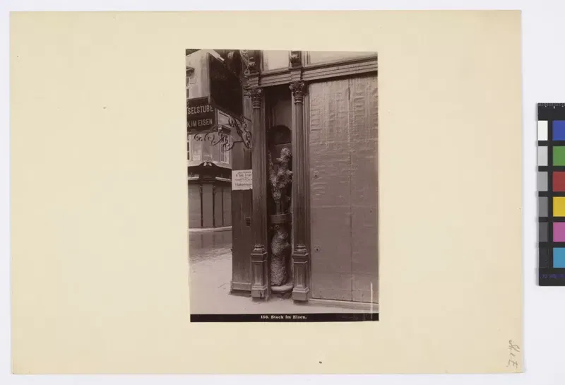Fotografie um 1880: der Stock im Eisen in einer schmalen Nische zwischen Holzpfeilern einer Hausfront, daneben ein Schild an der Wand