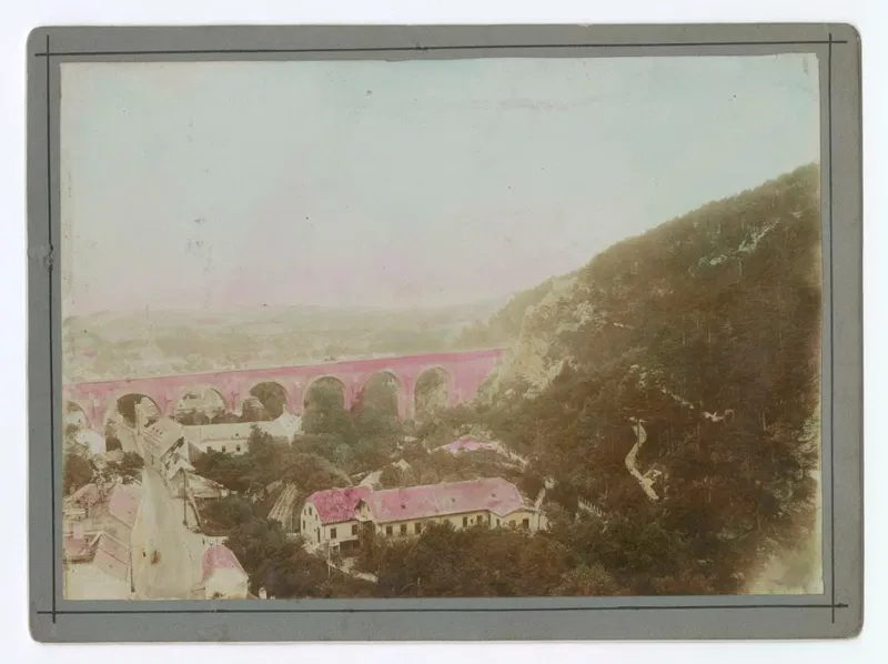 Kolorierte Fotografie: ein Aquädukt mit Bögen quer über das Tal hinter den Häusern von Mödling, rechts ein bewaldeter Hang