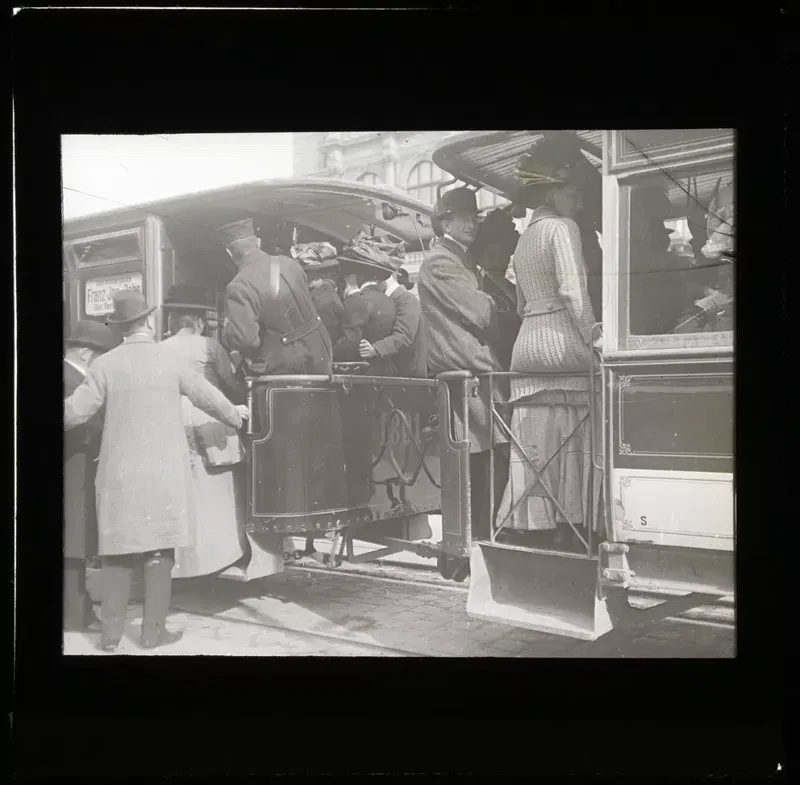 Fotografie 1905–1911: Fahrgäste mit Hüten drängen sich auf der hinteren Plattform einer Straßenbahn, unten ein Mann im langen hellen Mantel, rechts eine Frau im gestreiften Kleid