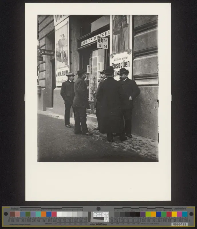 Fotografie um 1910: vier Männer in Mänteln und Hüten stehen vor dem Eingang einer Schenke mit Plakaten
