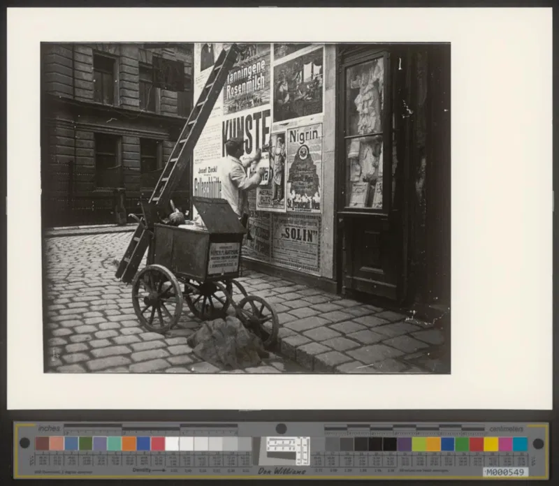 Fotografie um 1910: ein Mann klebt Plakate an eine Hauswand, davor ein Karren mit Leiter und Kübel auf Kopfsteinpflaster