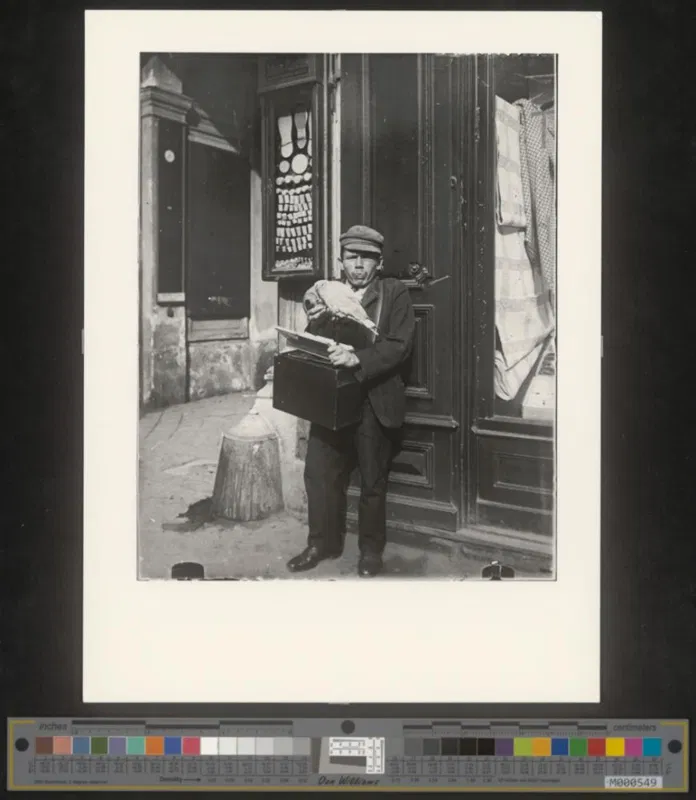 Fotografie um 1910: ein Mann mit Schirmkappe steht in einem Hauseingang und hält einen Kasten vor sich
