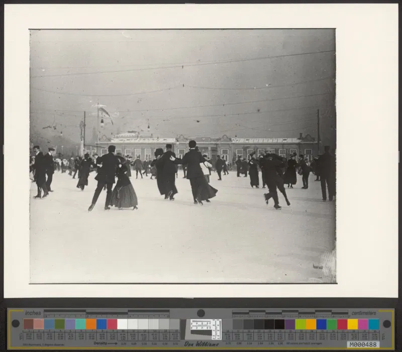 Fotografie um 1910: viele Paare in dunkler Kleidung laufen auf einer großen Eisfläche Schlittschuh, im Hintergrund ein langgestrecktes Gebäude