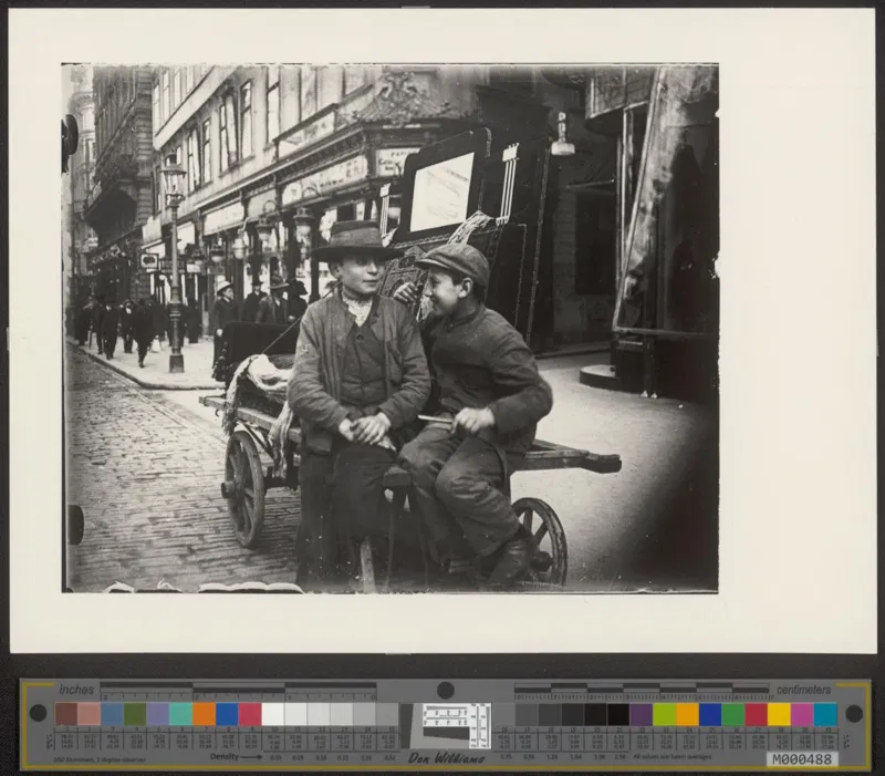 Fotografie um 1910: zwei Buben sitzen auf einem Handkarren in einer belebten Geschäftsstraße, einer mit Hut, einer mit Kappe