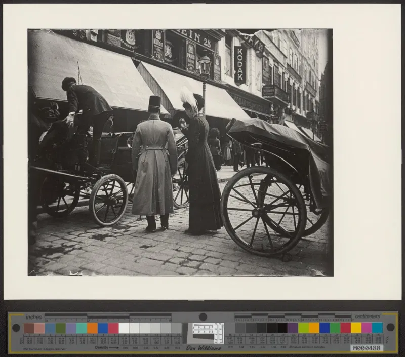 Fotografie um 1910: Kutschen in einer Geschäftsstraße mit Markisen, ein Mann im langen Mantel mit Zylinder und eine Frau im dunklen Kleid an einem Wagen