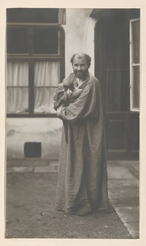 Fotografie 1911: Gustav Klimt im langen Malerkittel steht im Hof vor einem Haus und hält eine Katze im Arm