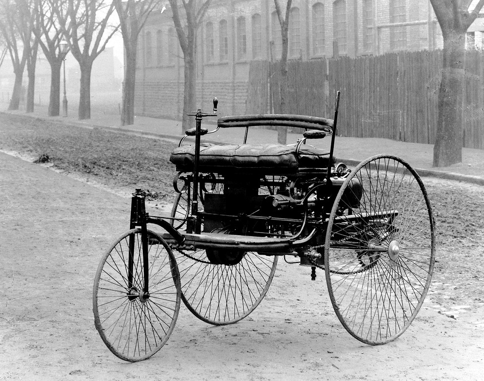 Historische Fotografie eines dreirädrigen Motorwagens mit großen Speichenrädern auf einer Straße.