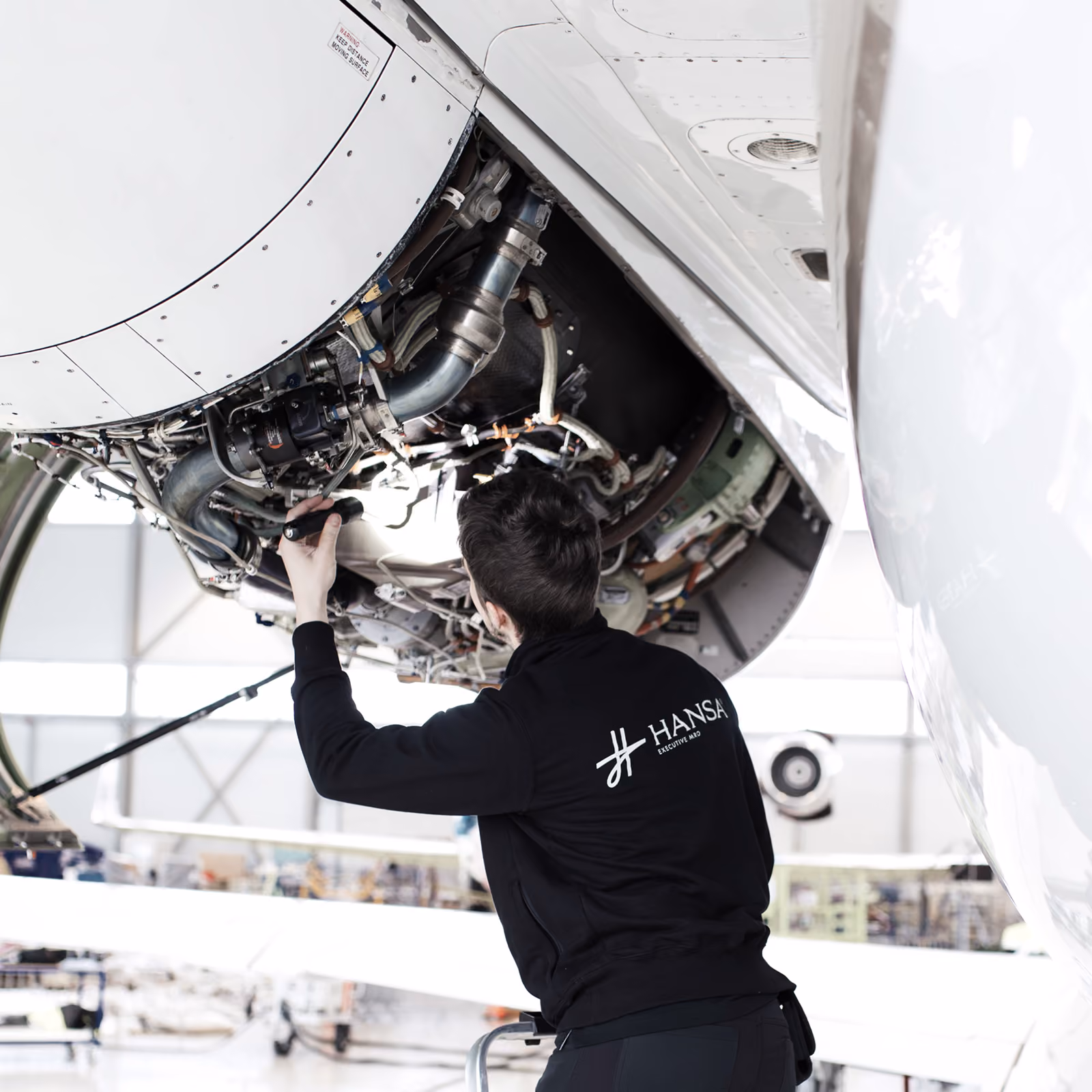 Line maintenance technician wearing a black jacket with Hansa Executive MRO logo inspects the internal components of a business jet engine in a hangar.