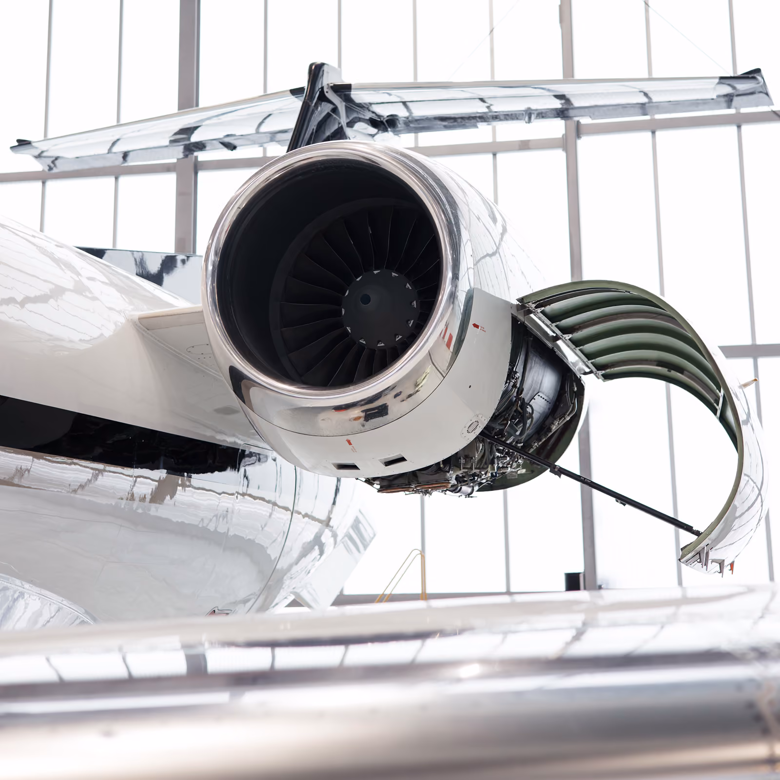 Close-up of an aircraft jet engine with its cowling open, mounted on the rear fuselage under the tail, inside Hansa Executive MRO hangar with large windows.