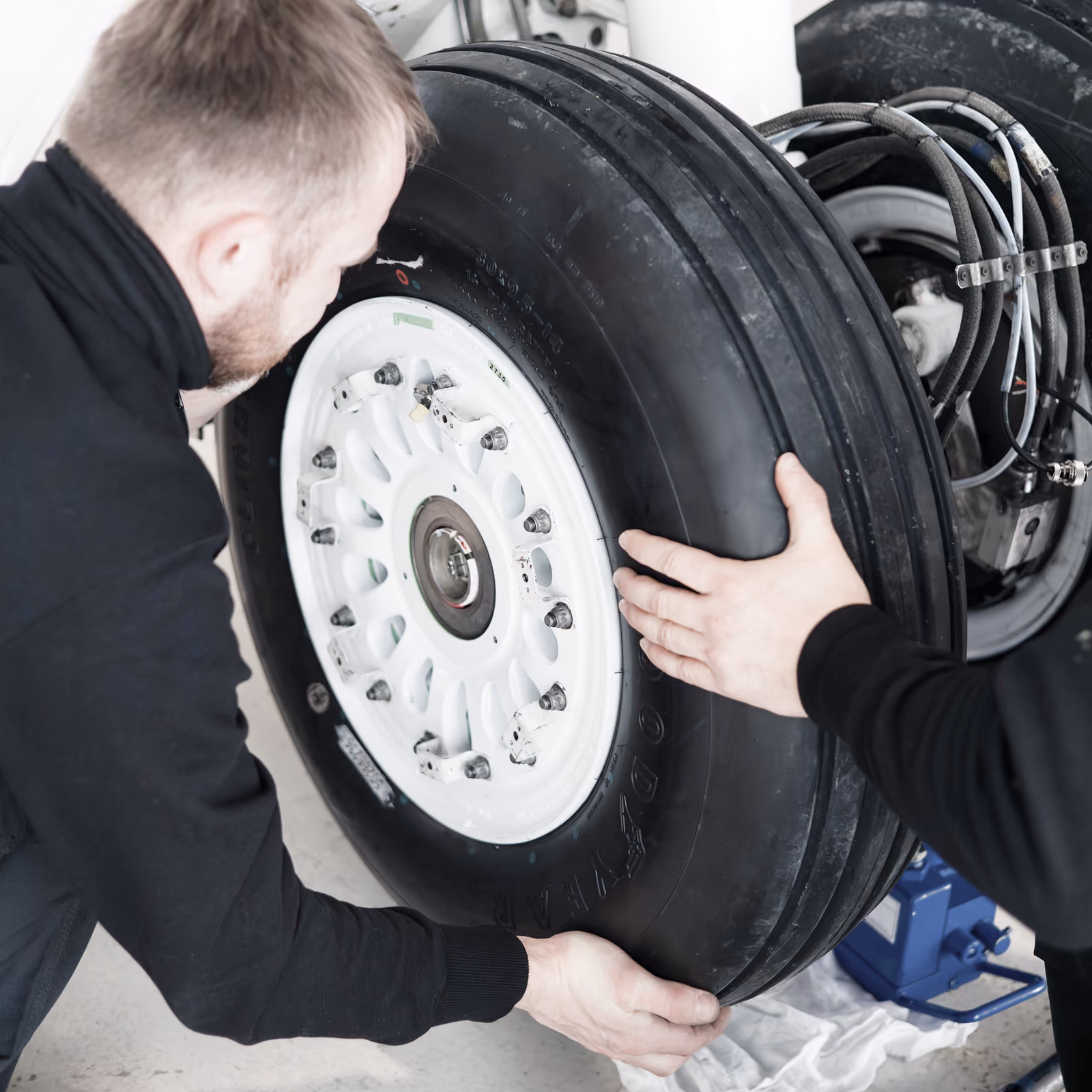 Two men in black Hansa Executive MRO shirts handling and inspecting a large business jet landing gear tire with a white rim, mounted on an aircraft wheel assembly.